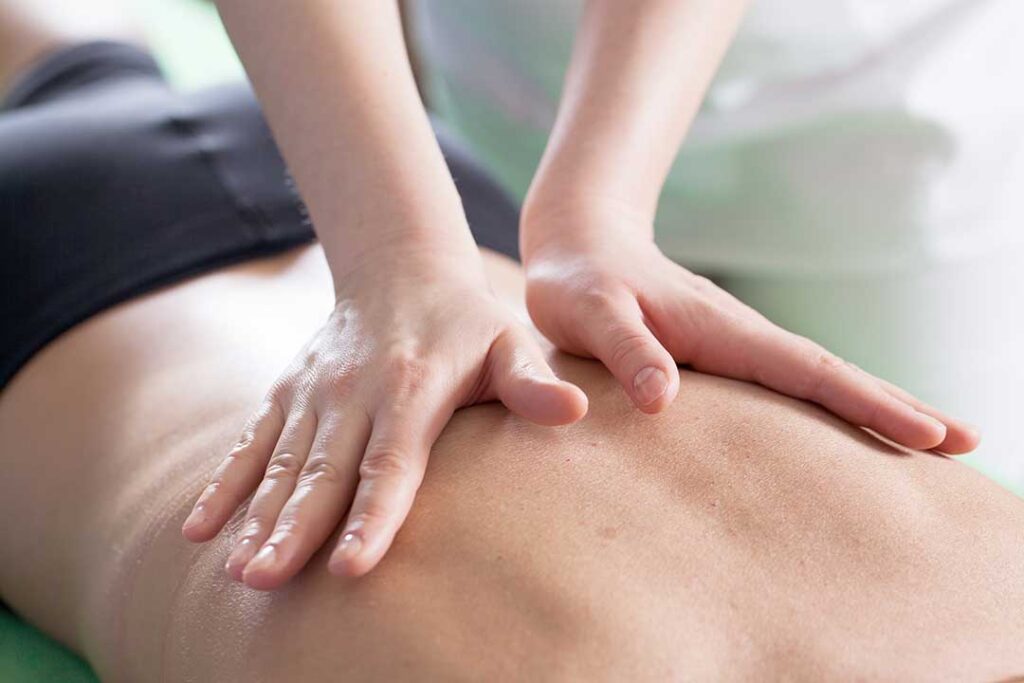A person receives gentle back massage while a clinician discusses spinal movement beside a treatment table.