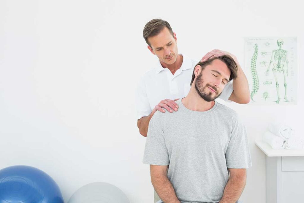 Person gently pressing a tender upper-back muscle while seated near a window.